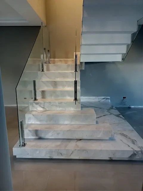 Escada em pedra clara de veios cinza, com guarda-corpo de vidro